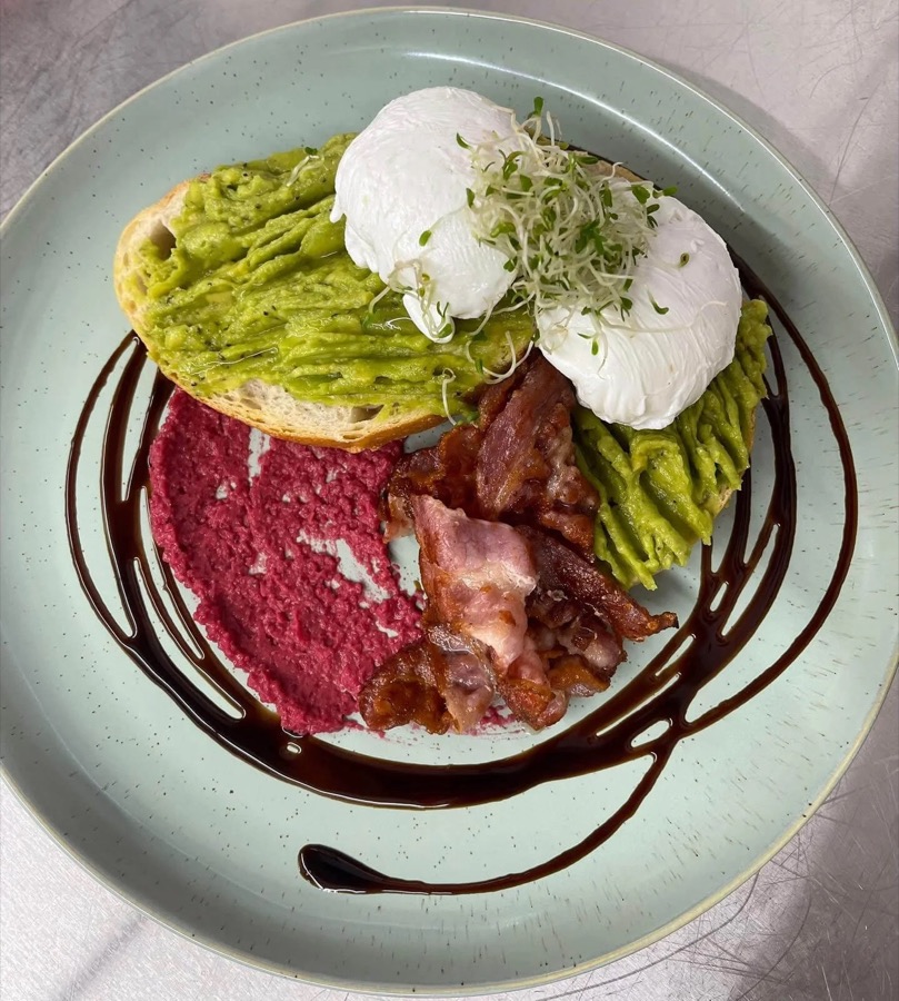 Smashed avocado on sourdough with two poached eggs, bacon and a balsamic swirl on a sage plate